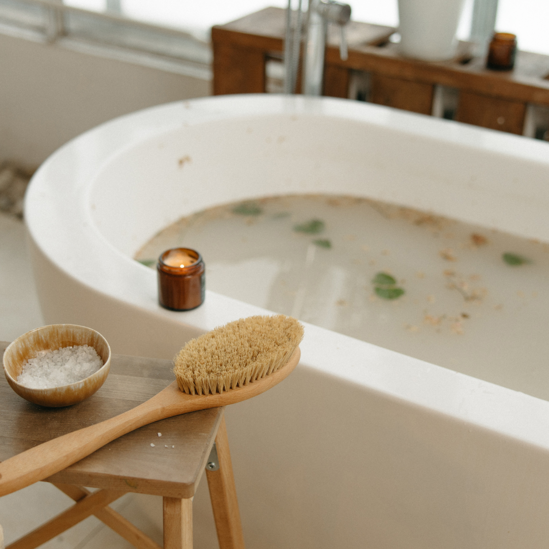 Bathtub filled with warm water and floating herbs, styled with a wooden bath brush, salt bowl, and candle in a calm bathroom setting.