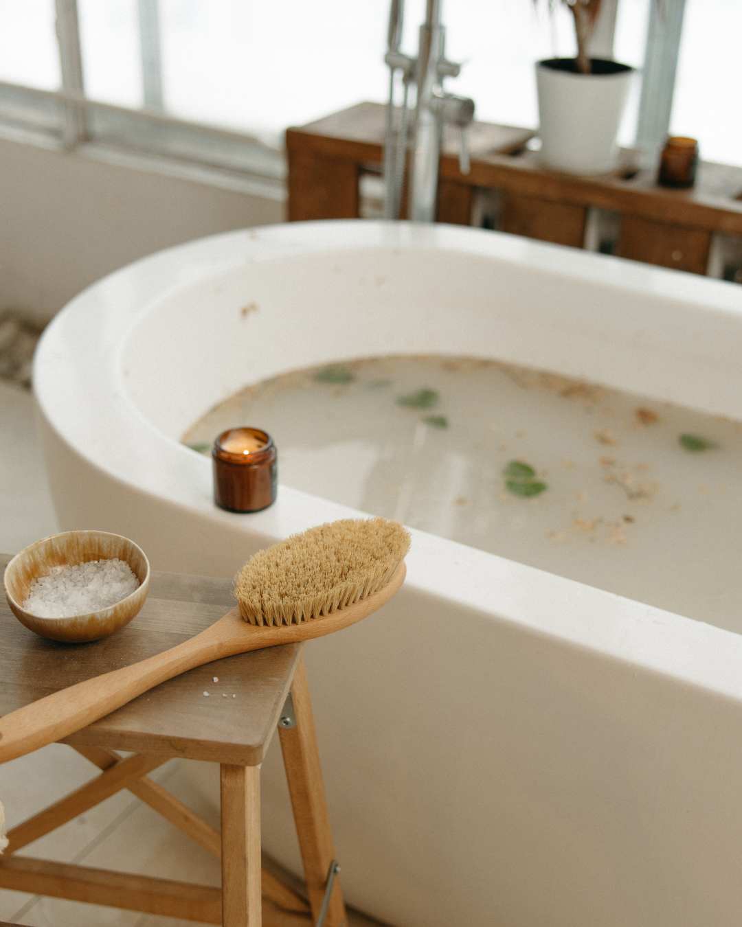 Bathtub filled with warm water and floating herbs, styled with a wooden bath brush, salt bowl, and candle in a calm bathroom setting.